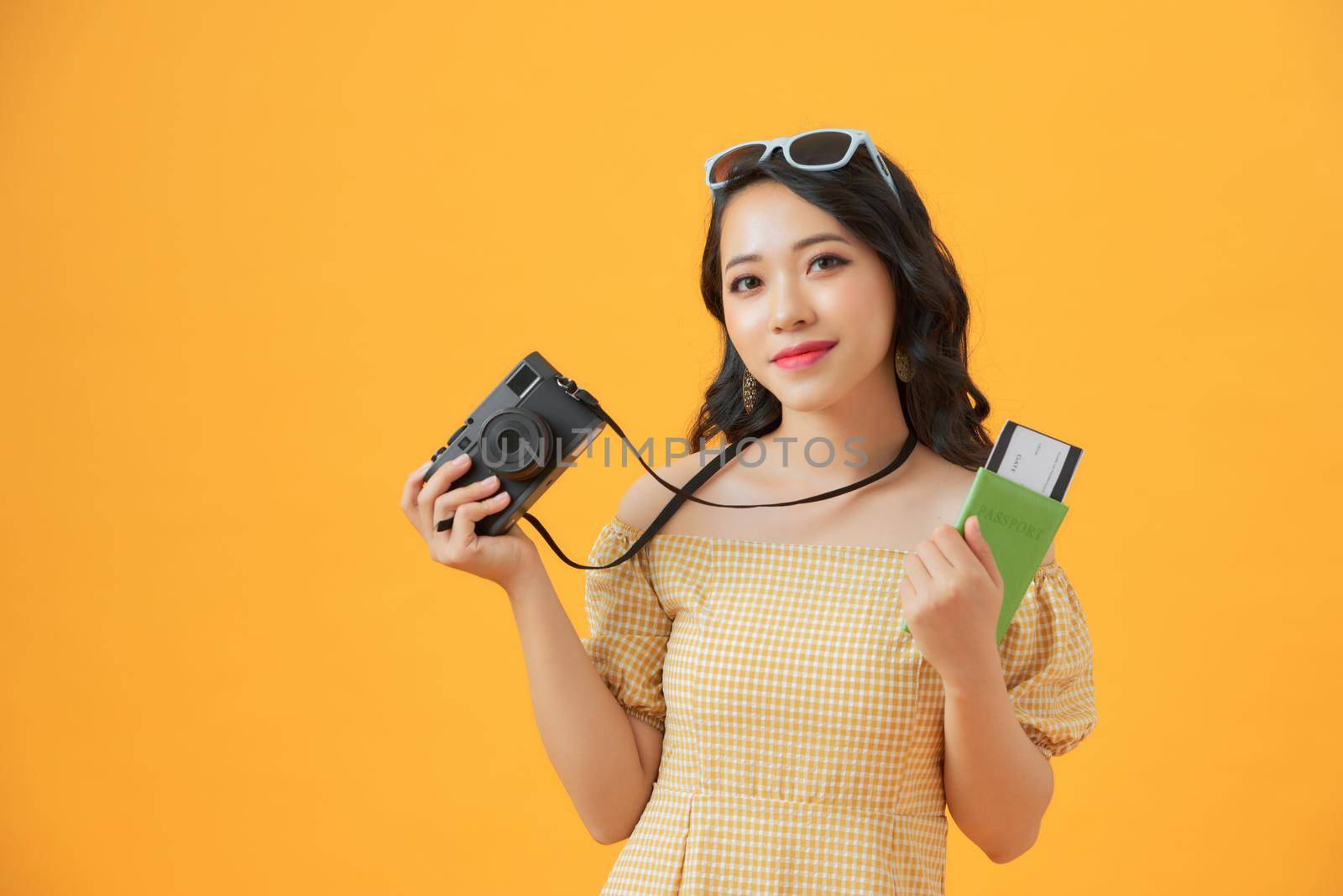 Portrait of a happy young woman holding camera and showing passport while standing isolated over yellow background