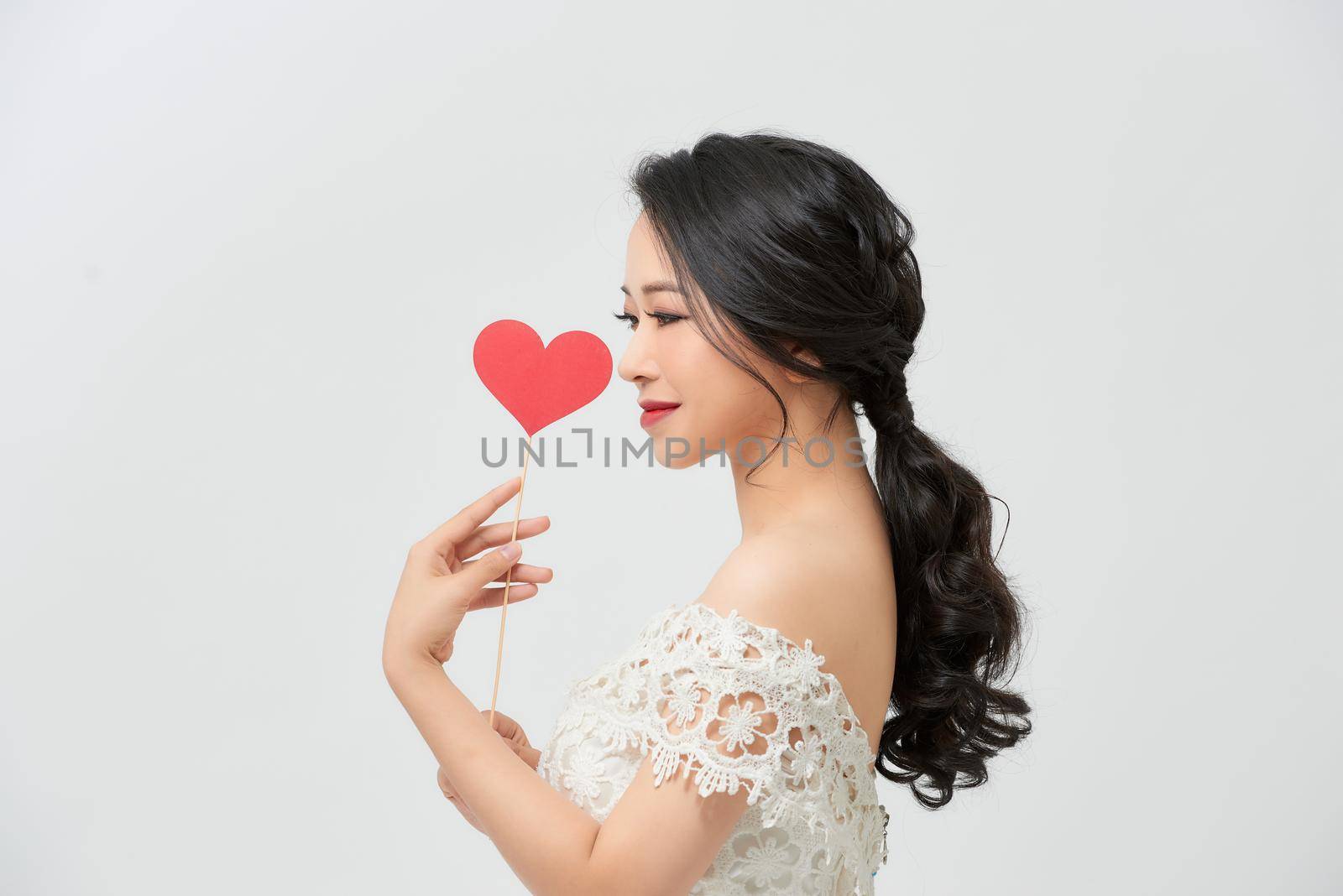 Elegant asian woman in white dress holding red heart on stick with smile