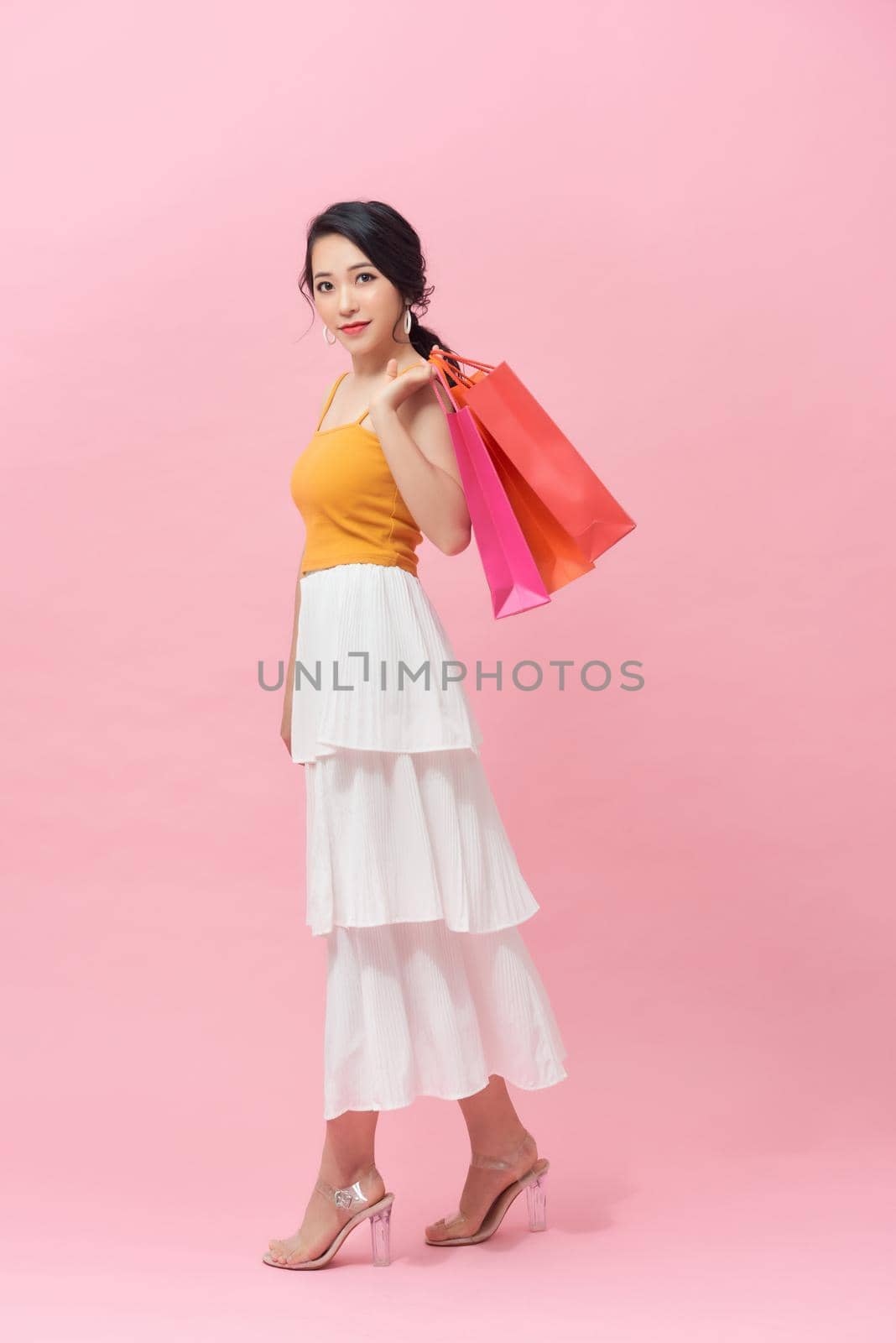 full length view of cheerful, fashionable woman holding colorfull shopping bags and smiling on pink background by makidotvn