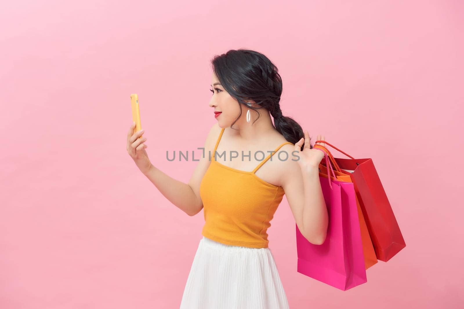 Happy Asian woman holding bags with purchases, smiling while looking at phone in shopping center. 
