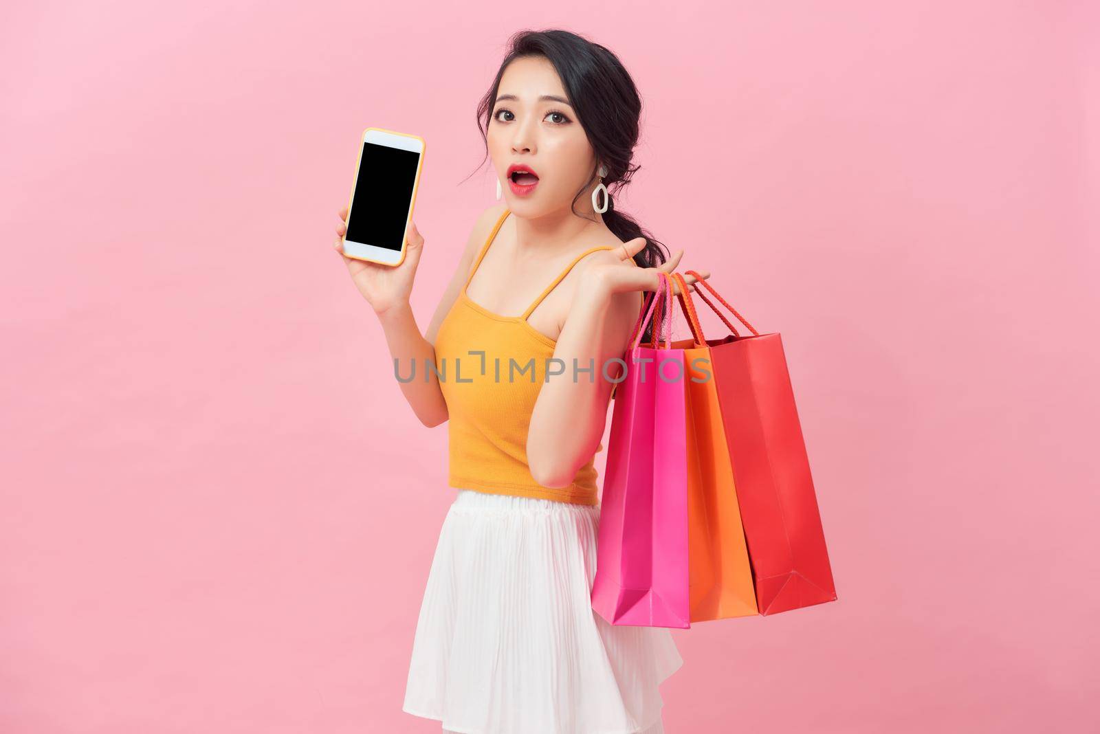 Photo of shocked woman 20s in dress looking at smartphone in hand with surprise while holding shopping bags isolated over pink background by makidotvn