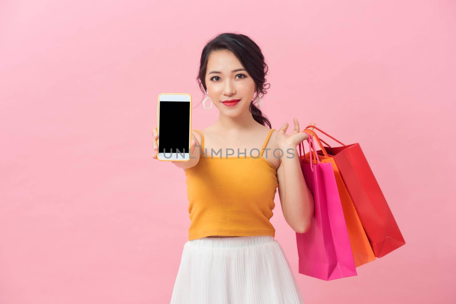 Portrait of a pretty excited girl with colorful shopping bags showing blank screen mobile phone over pink background. by makidotvn