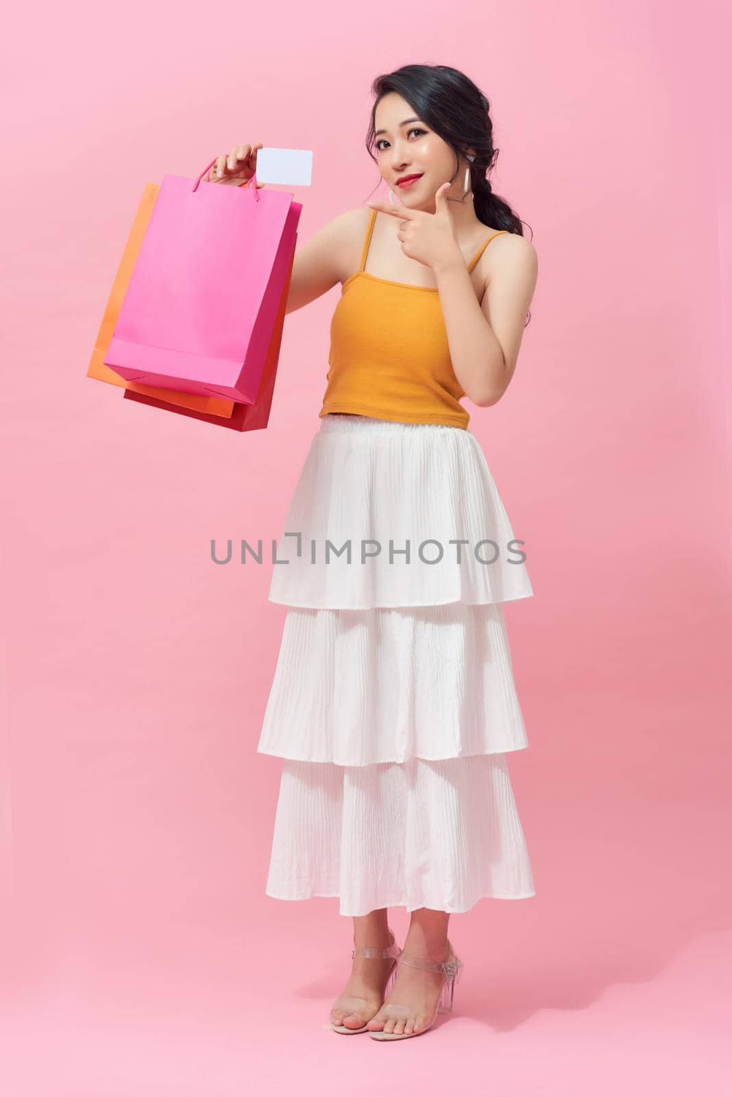 Full length of excited Asian woman with colorful shopping bag while standing over pink background. by makidotvn