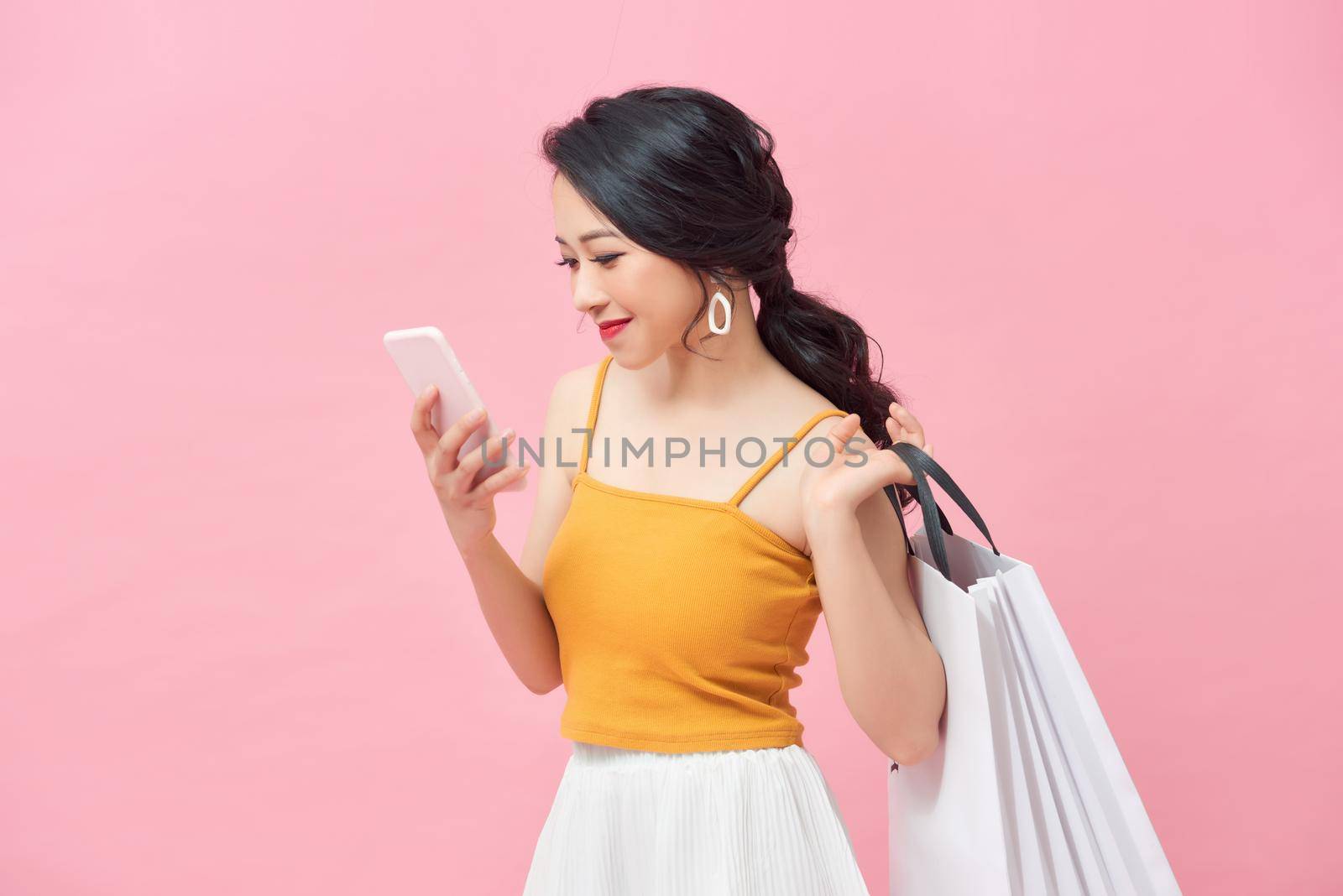 Beautiful girl is holding shopping bags, using a smart phone over pink background. by makidotvn