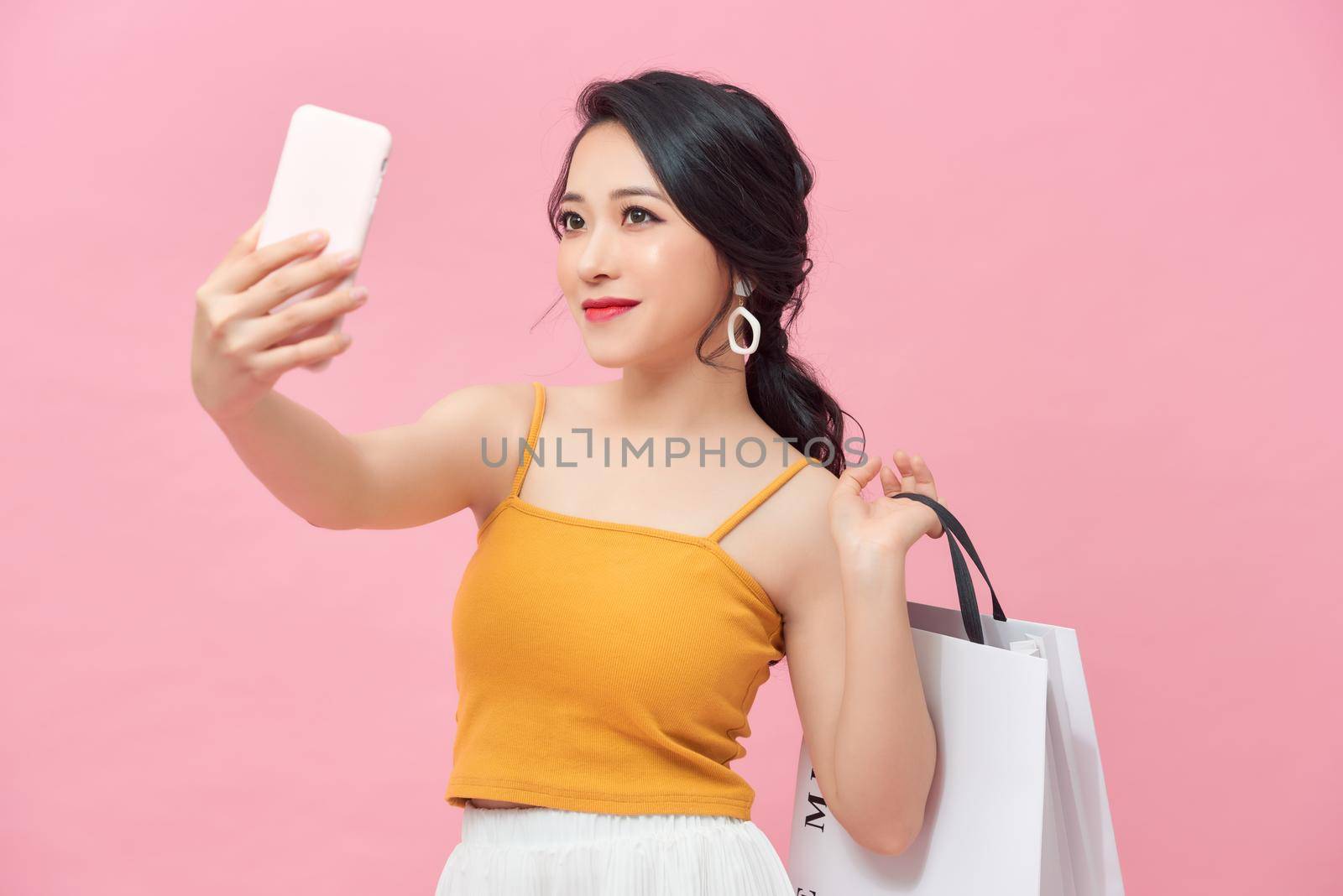 Attractive young Asian woman selfie while holding shopping bag over pink background.