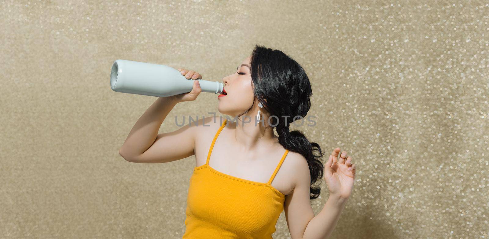 Attractive young Asian woman drinking wine wit bottle over gold background.