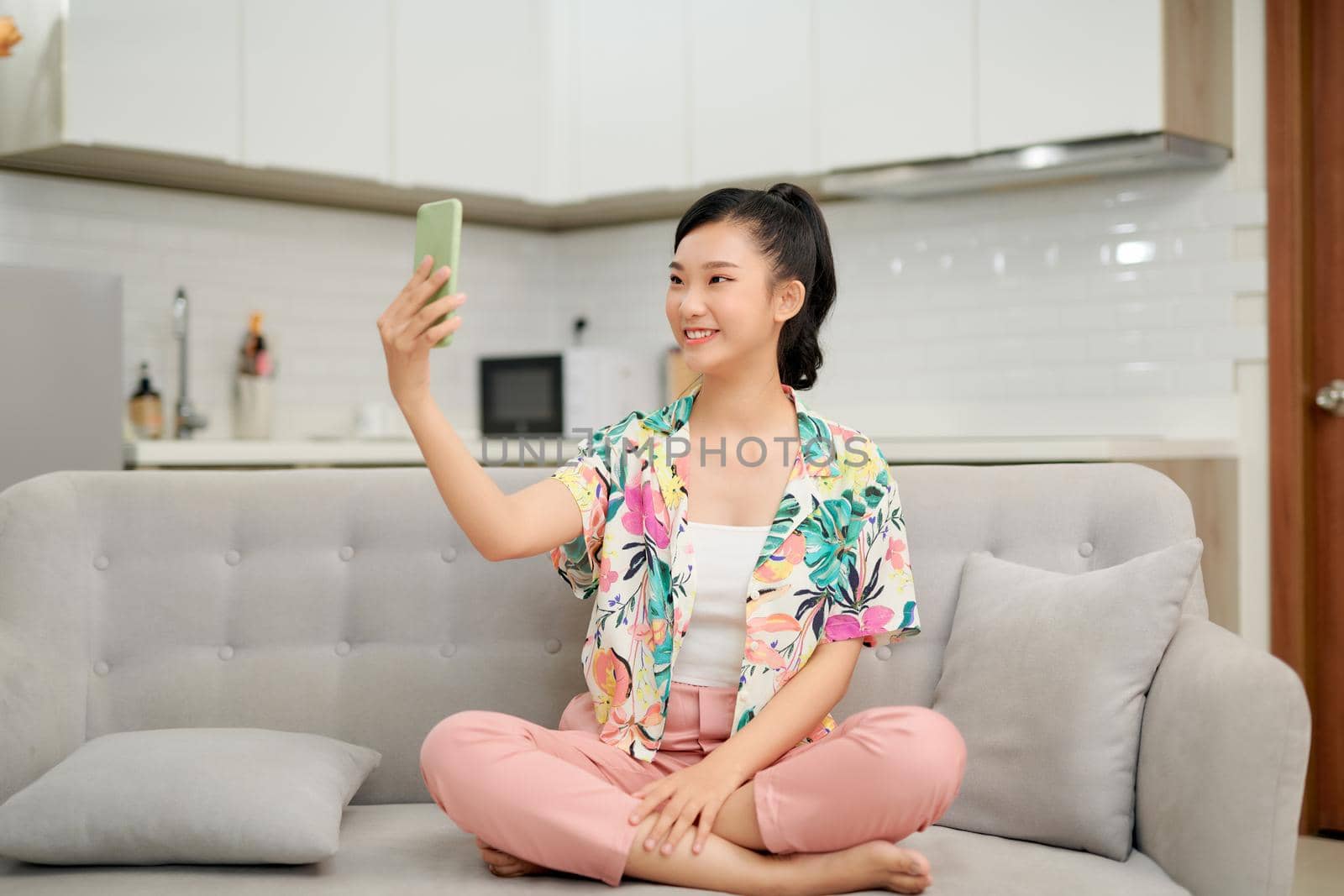 Pretty Young Asian Woman sits on sofa hand holding smartphone taking selfie