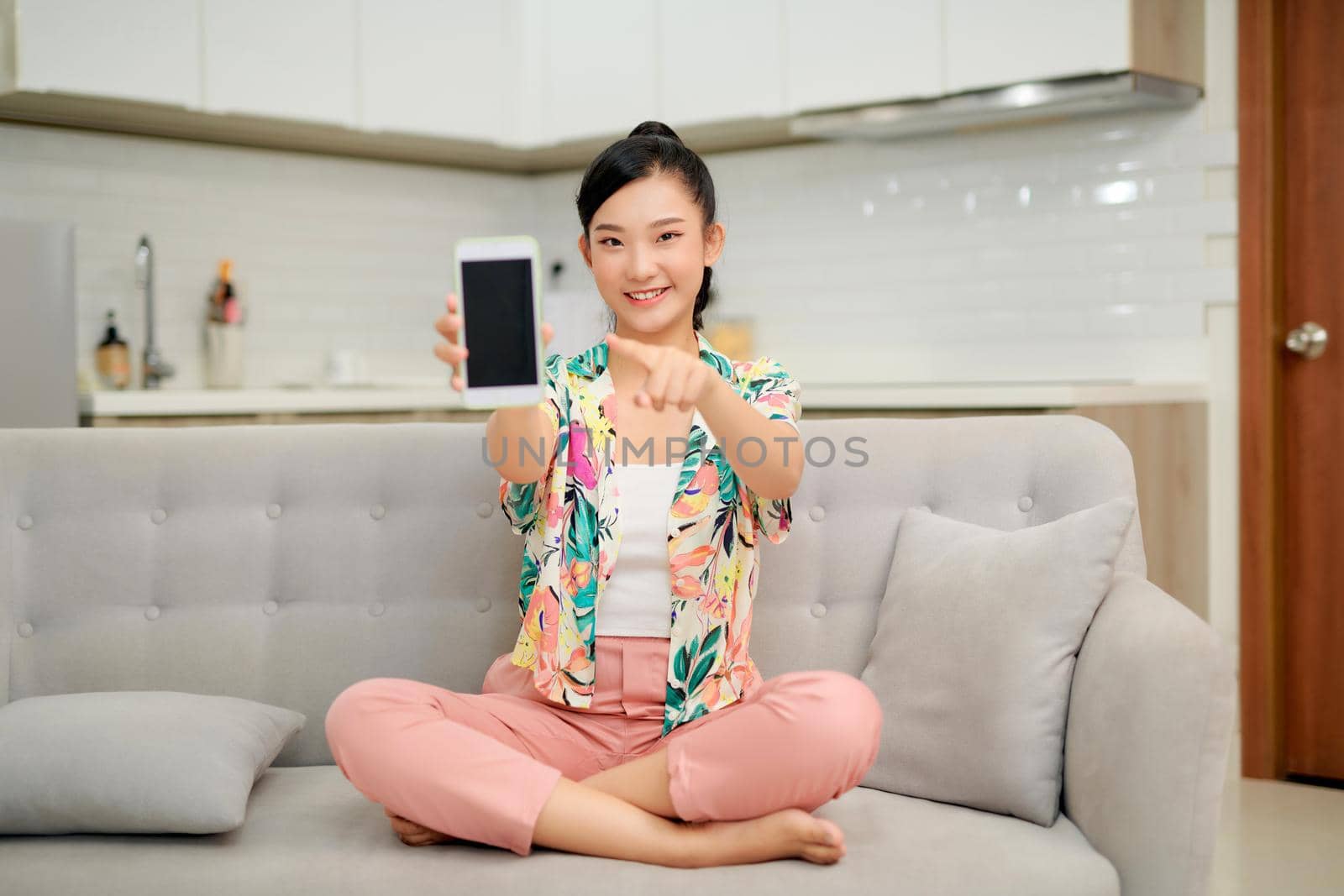 Smiling young woman sitting on couch at home, pointing index finger on mobile phone with blank empty screen