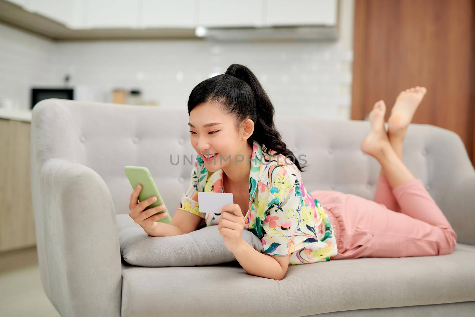 Young smiling Asian woman using smartphone buying online shopping by credit card while lying on sofa at home.