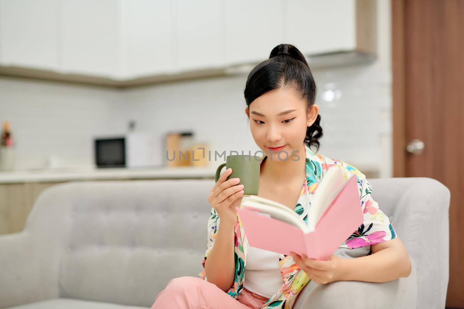 Young beautiful woman sitting on the sofa reading a book enjoying her tea in living room at home