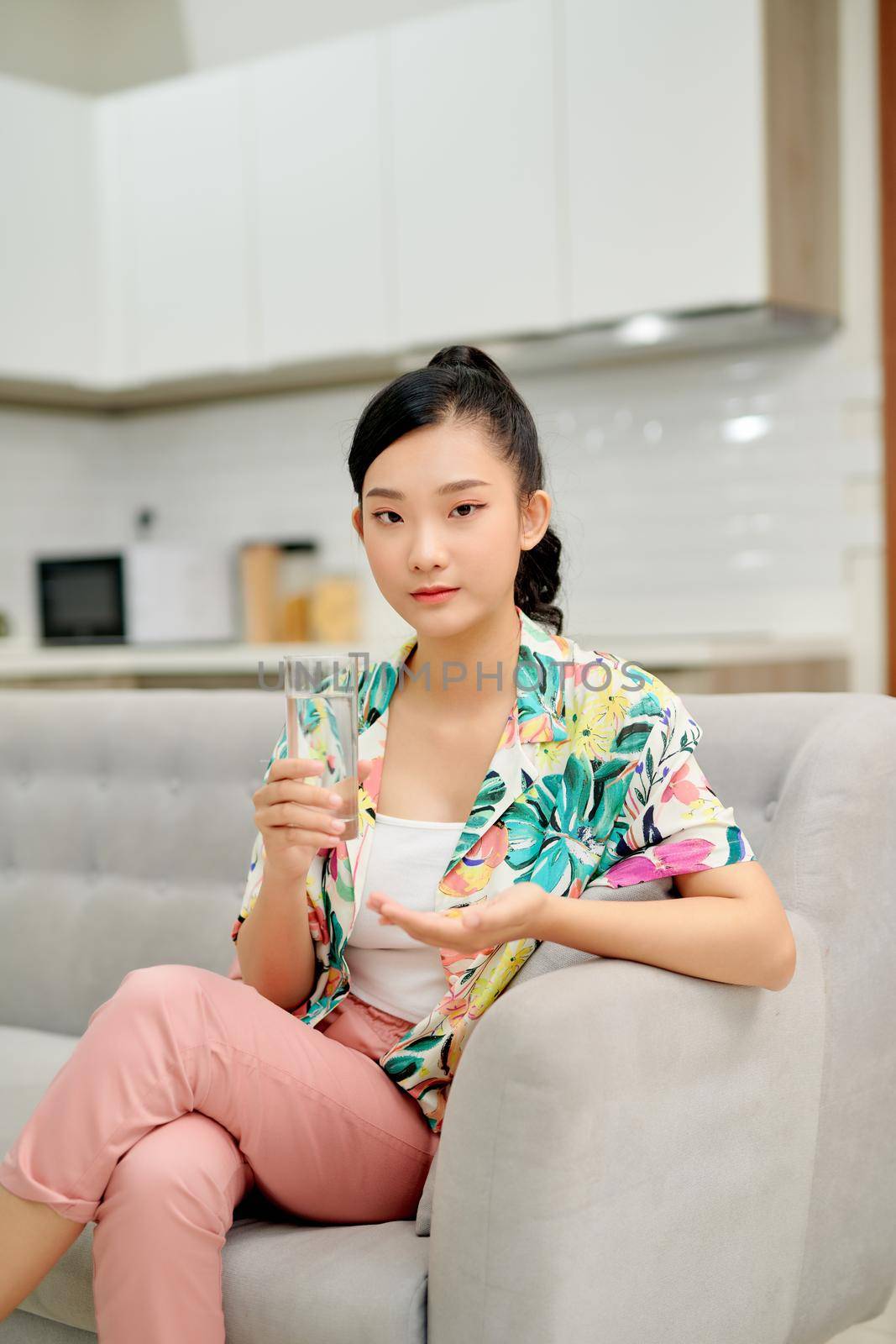 Happy woman taking vitamin pill holding glass sitting on a couch at home