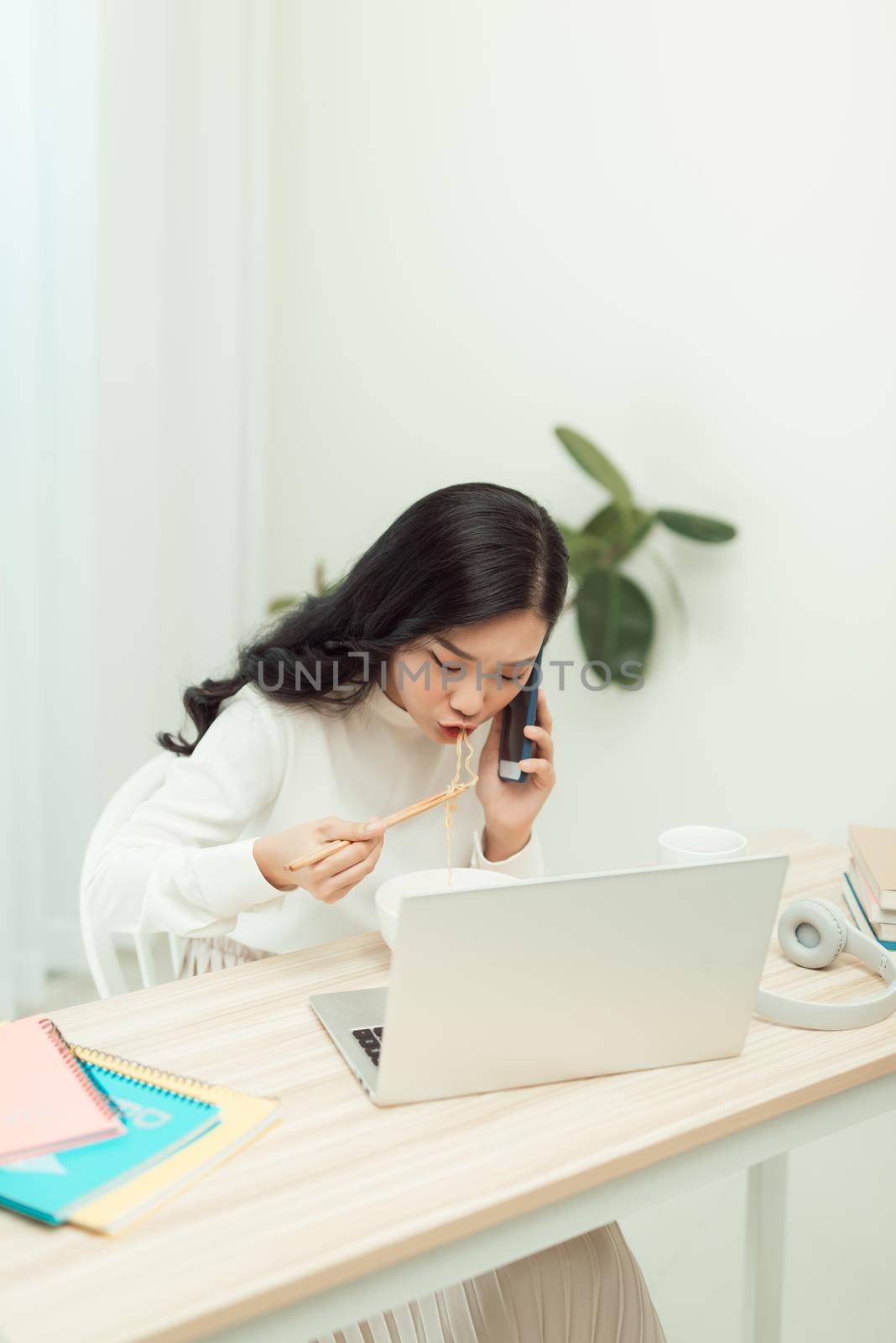 Confident smiling attractive young asian woman entrepreneur having lunch break while sitting at the office desk and using mobile phone by makidotvn