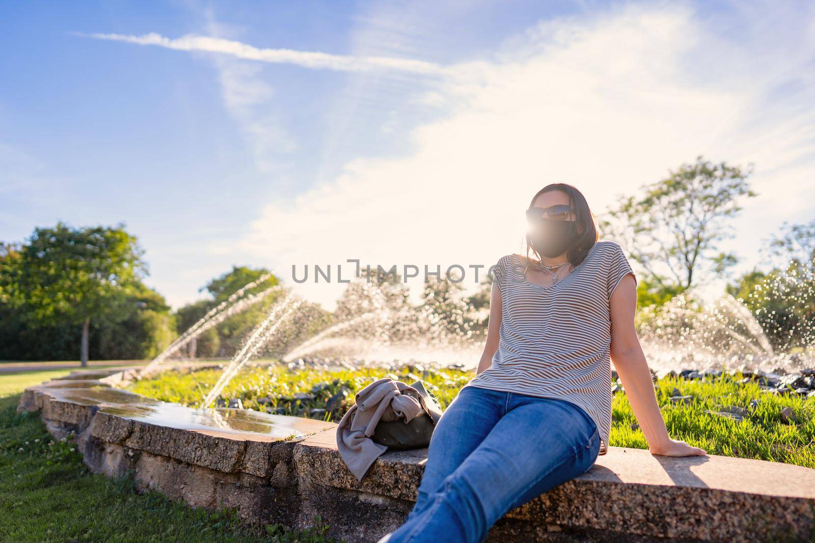 Young woman alone in the park sitting on a fountain edge in protective mask against Coronavirus for social distancing with blue sky, white clouds and splashing water among the reflections of the sun by robbyfontanesi