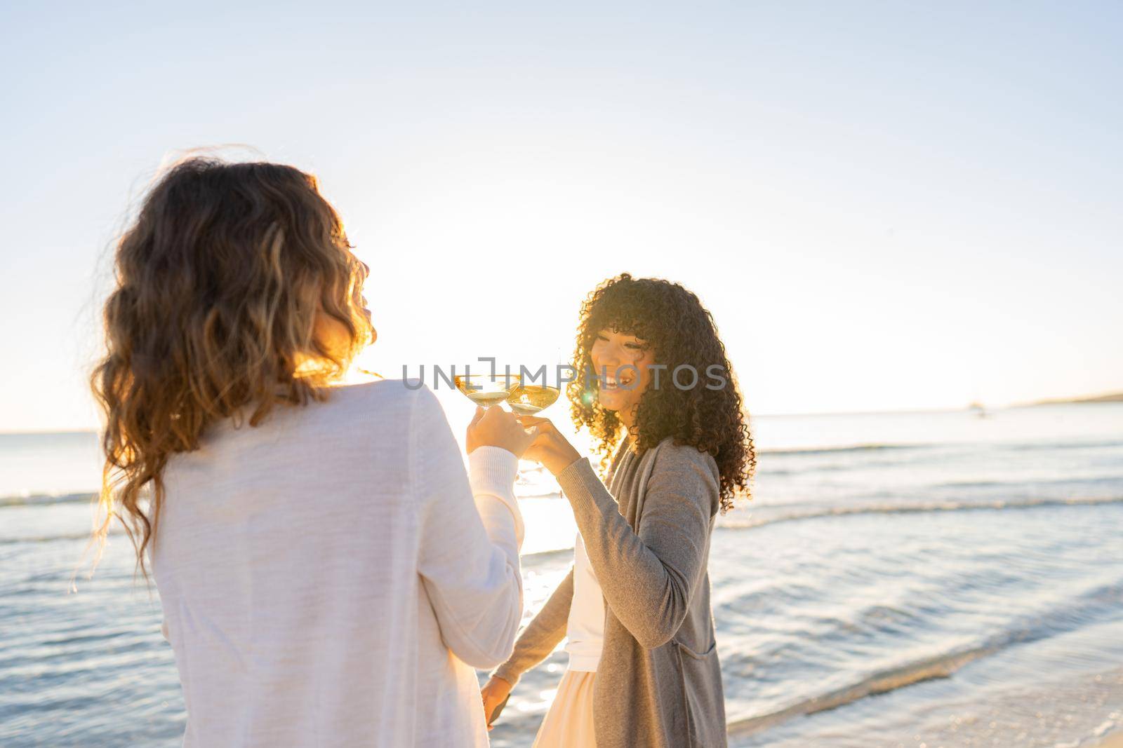 Two beautiful trendy young lesbian girlfriends laugh toasting with white wine at sunset or sunrise with sun reflections running through the crystal glasses. Stylish women having fun on beach