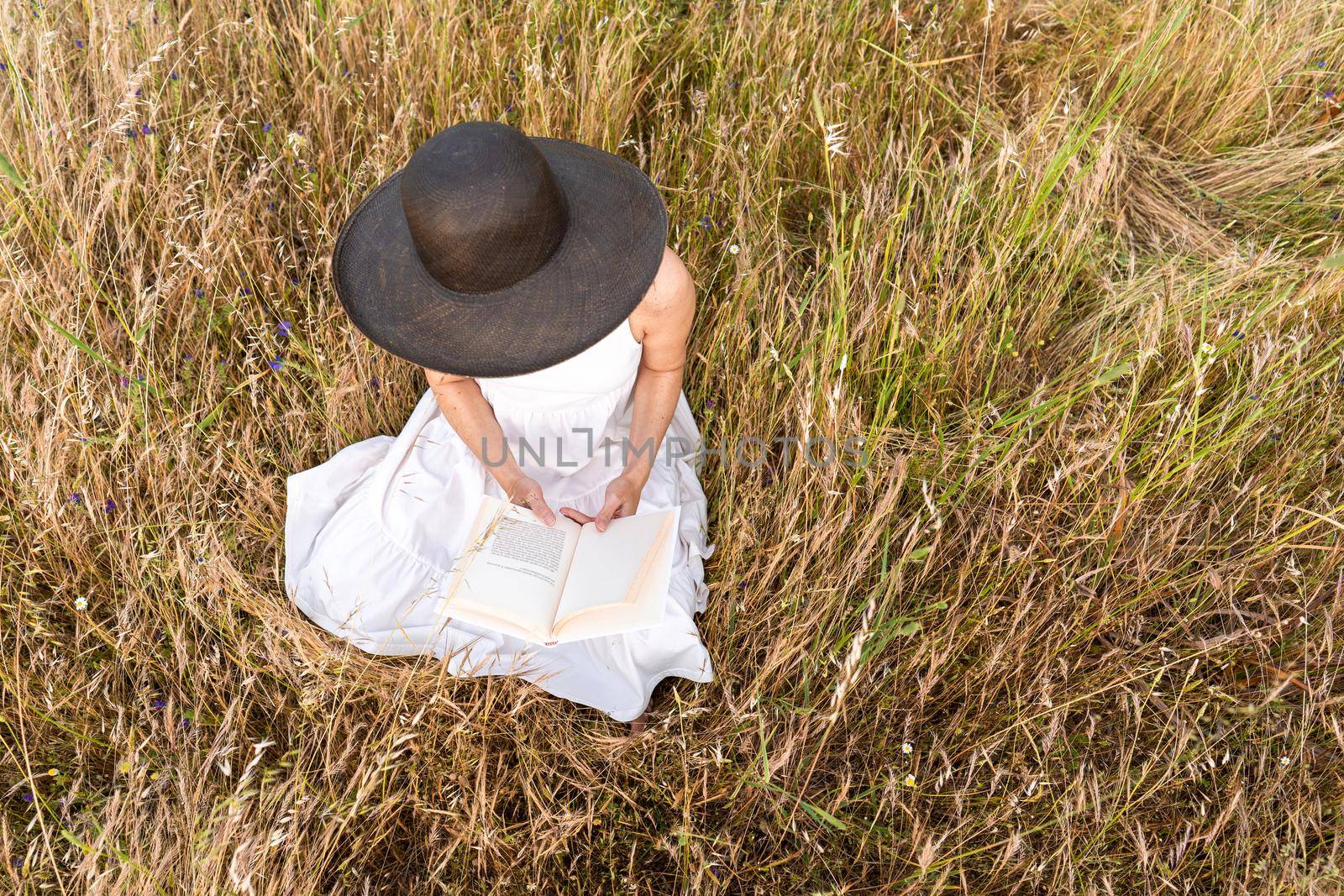 Romantic dreamy top view scene of unrecognizable woman sitting in a field of wheat and tall yellow grass wearing a dark wide-brimmed hat holding a book. Boho girl reading preferred romance story tale