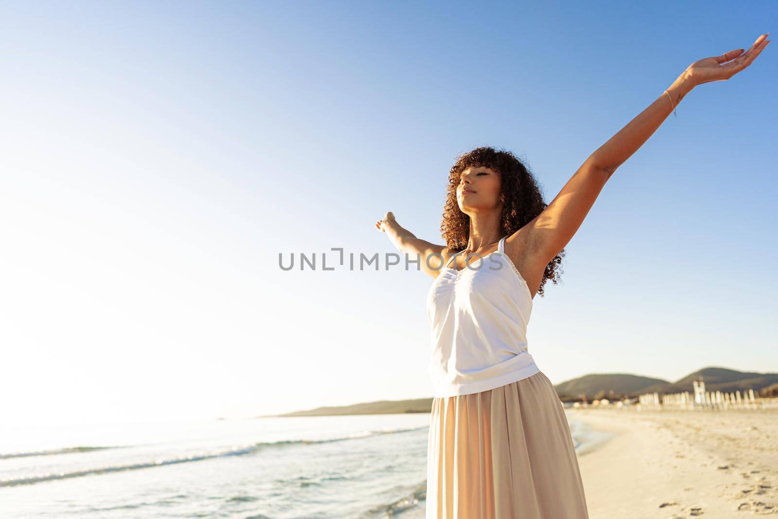 Young beautiful curly black hispanic woman in boho dress open arms inspired from nature at sunset or sunrise. African-American brunette girl experiencing ocean sea energy at beach resort in vacations by robbyfontanesi