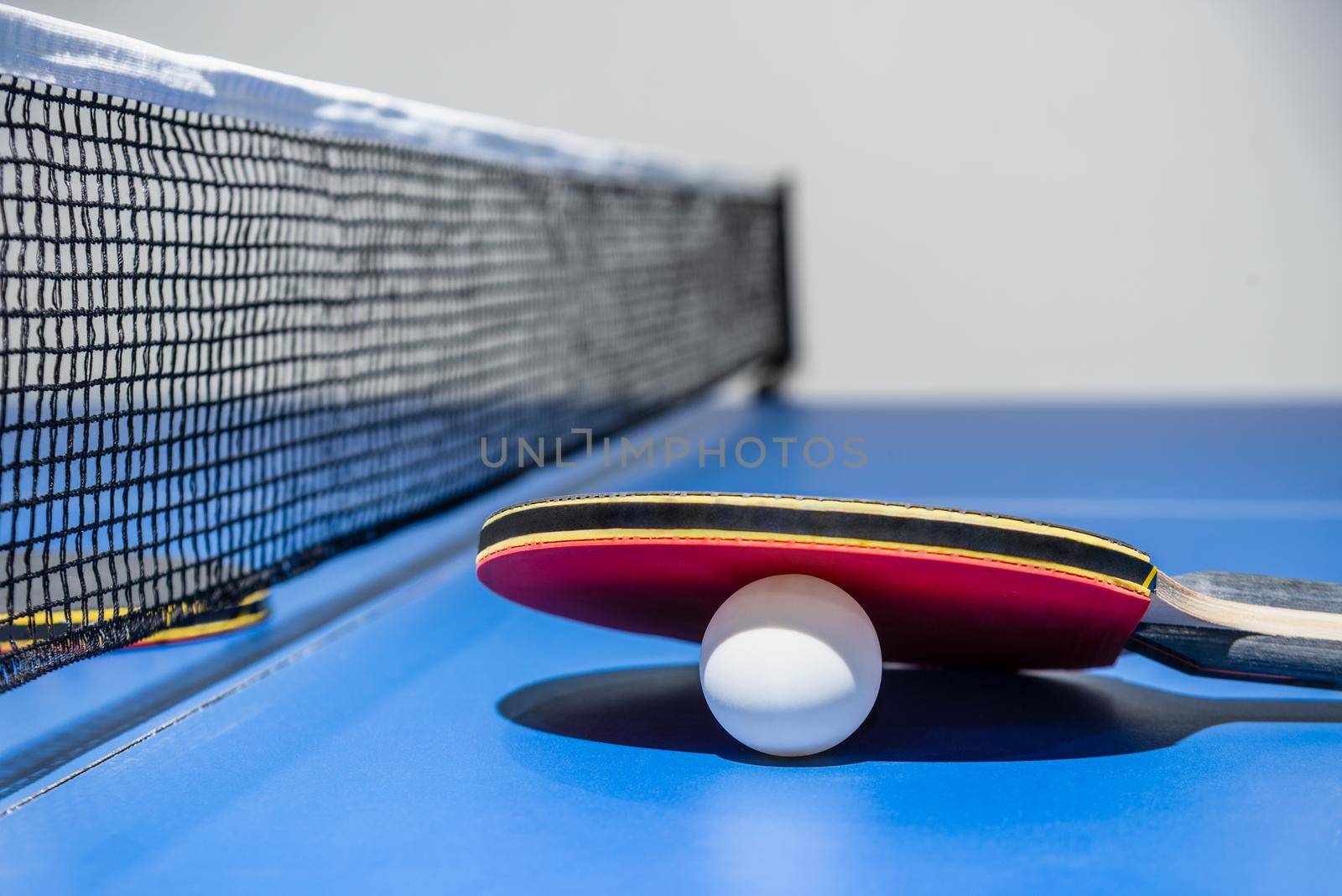 Closeup red table tennis racket and a white ball on the blue ping pong table with a black net, Table tennis paddle is a sports competition equipment indoor activity and exercise for concept background