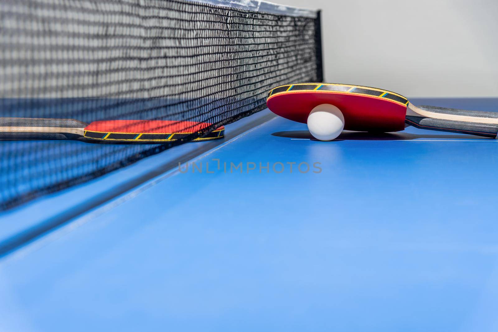 Red table tennis racket and a white ball on the blue ping pong table with a black net, Two table tennis paddle is a sports competition equipment indoor activity and exercise for concept background