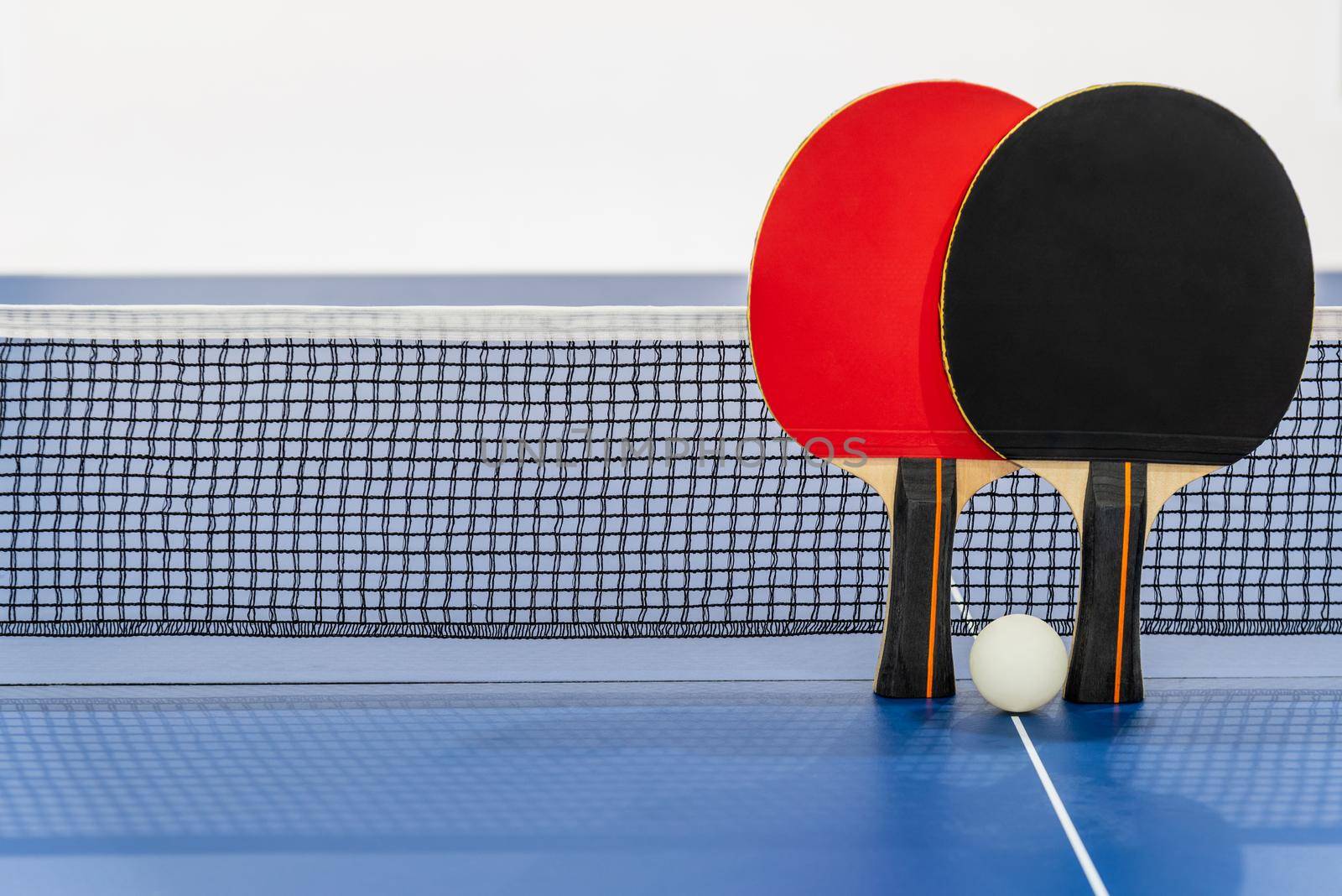 Black and red table tennis racket and a white ball on the blue ping pong table with a net, Two table tennis paddle is a sports competition equipment indoor activity and exercise for background concept