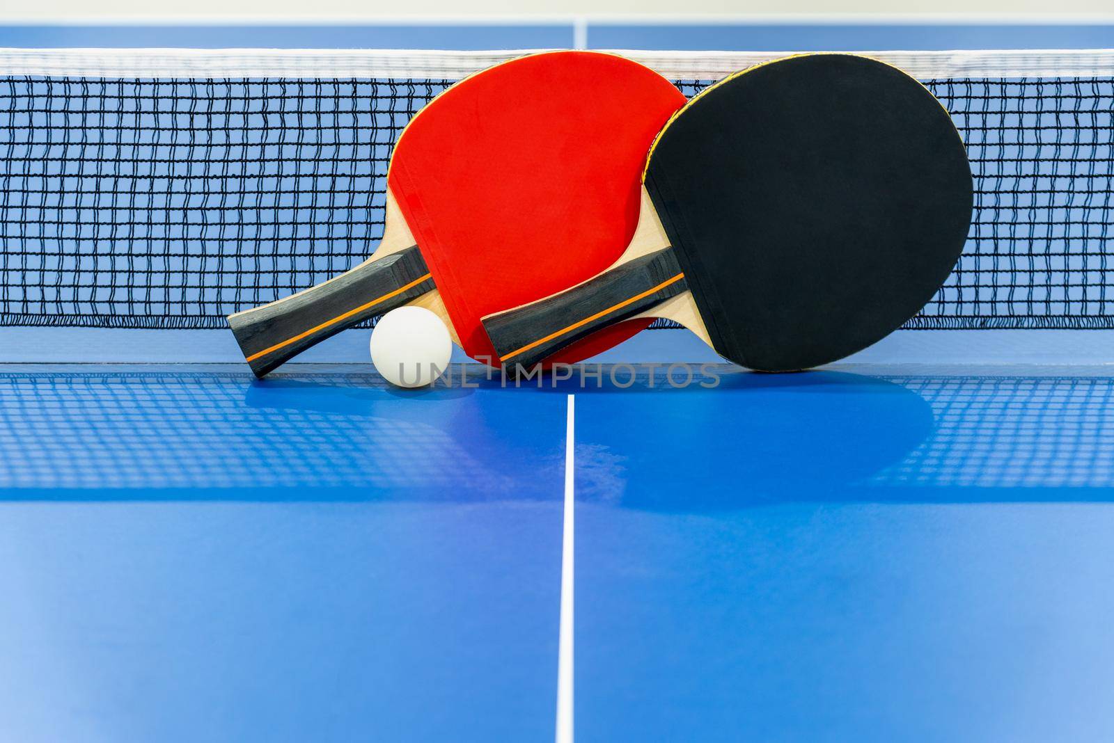 Black and red table tennis racket and a white ball on the blue ping pong table with a net, Two table tennis paddle is a sports competition equipment indoor activity and exercise for background concept