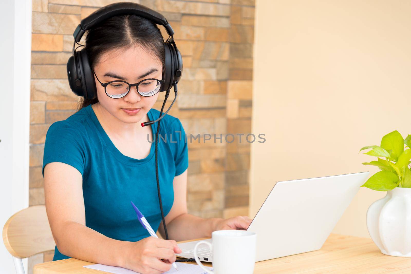 Asian teenage girl student with a headset is taking note learned online from school in a notebook using a laptop computer. Distance education from the class of the university by a video call from home