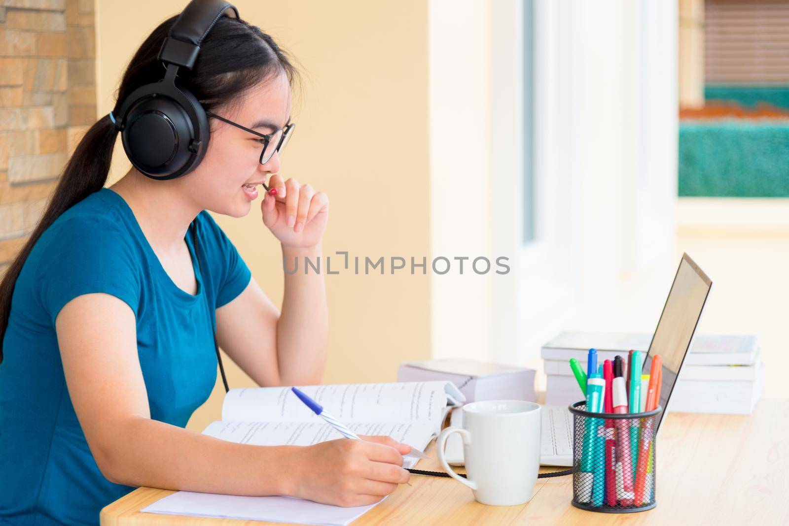 Asian teenage girl student with a headset and eyeglasses looks at laptop computer online learning from school and speak into a microphone. Distance education class of the college video call at a home