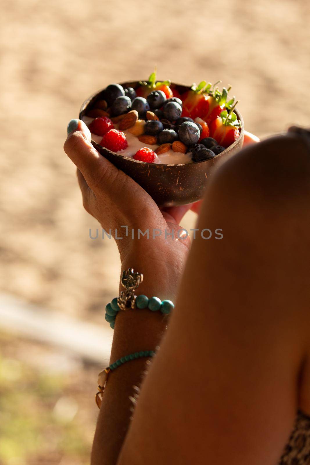 Woman holding a delicious smoothy breakfast bowl filled with fresh summer fruits, yoghurt and muesli in a coconut bowl on a beach in the morning.