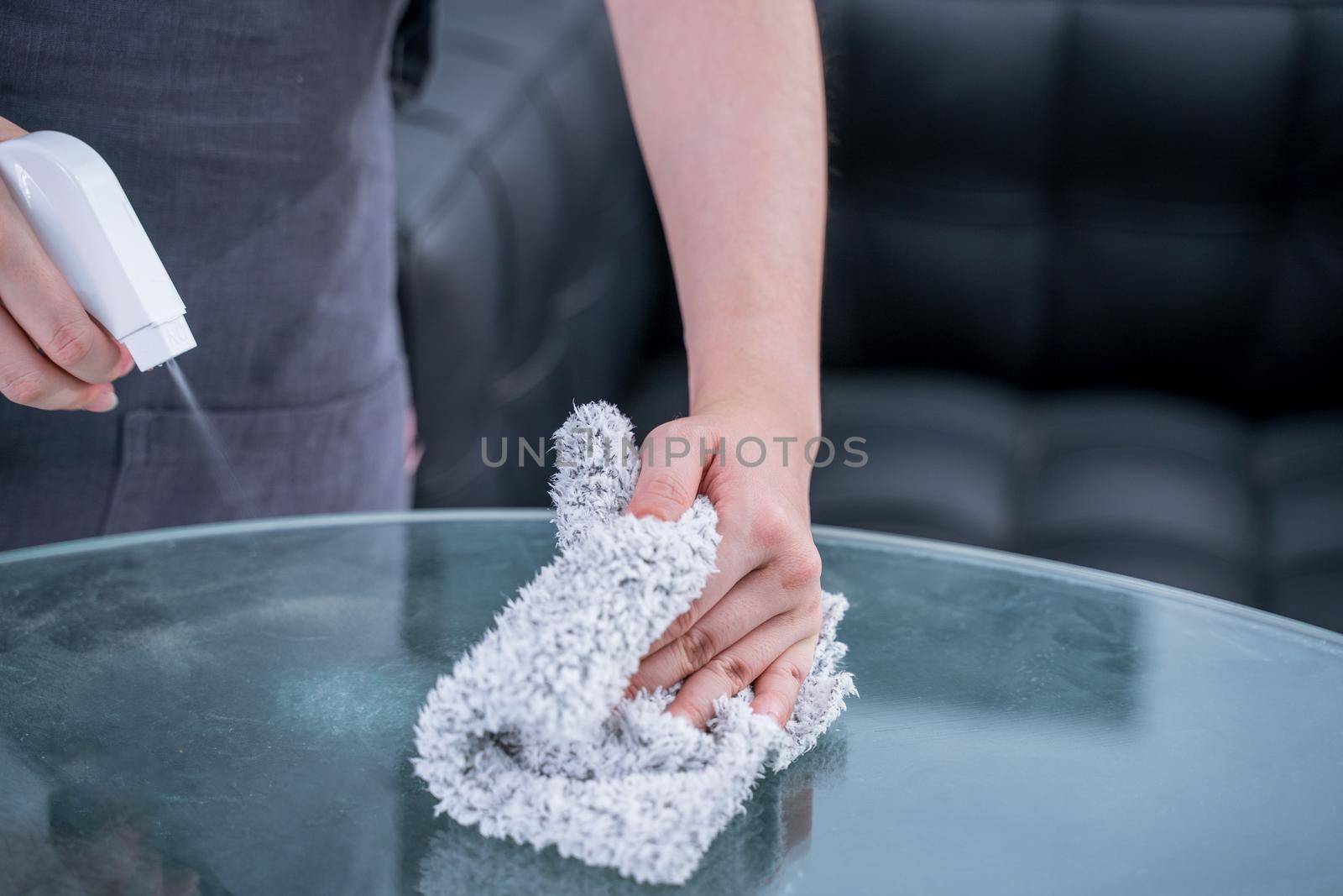 Young woman housekeeper in apron is cleaning, wiping down table surface with wet rag, spraying bottle cleaner to prevent disease spreading.