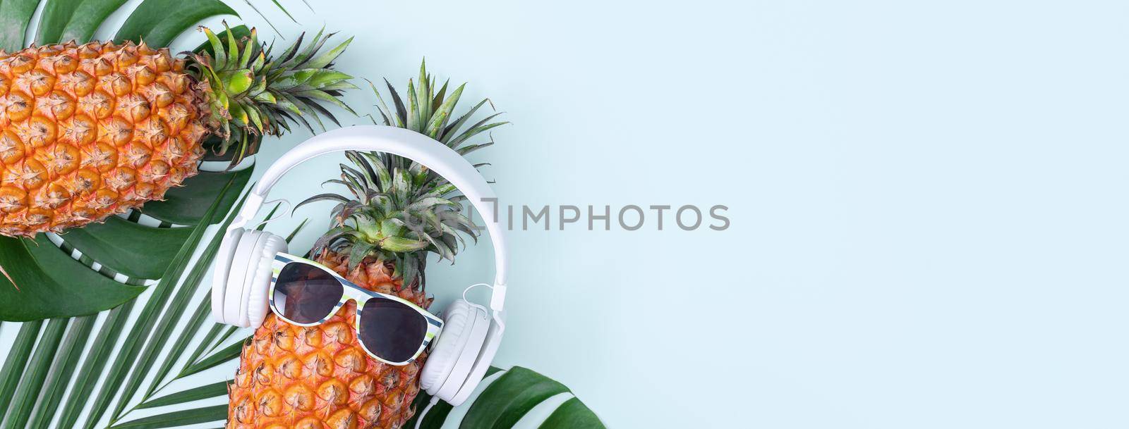 Funny pineapple wearing white headphone, concept of listening music, isolated on colored background with tropical palm leaves, top view, flat lay design.