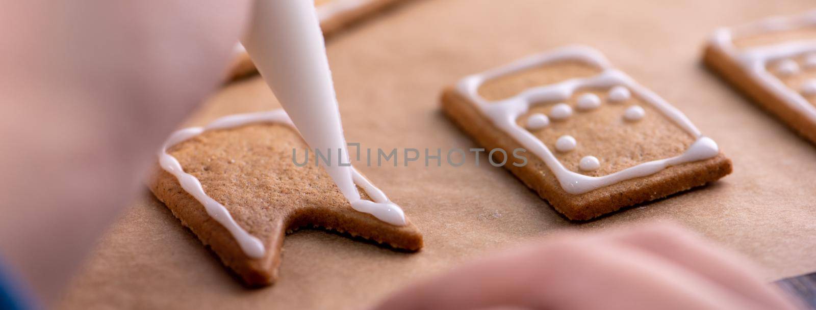 Young woman is decorating Christmas Gingerbread House cookies biscuit at home with frosting topping in icing bag, close up, lifestyle.