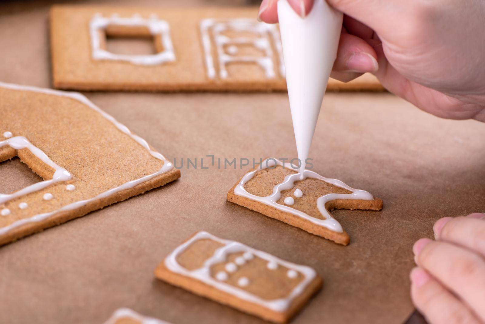 Young woman is decorating Christmas Gingerbread House cookies biscuit at home with frosting topping in icing bag, close up, lifestyle.
