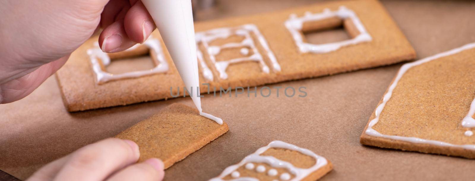 Young woman is decorating Christmas Gingerbread House cookies biscuit at home with frosting topping in icing bag, close up, lifestyle.