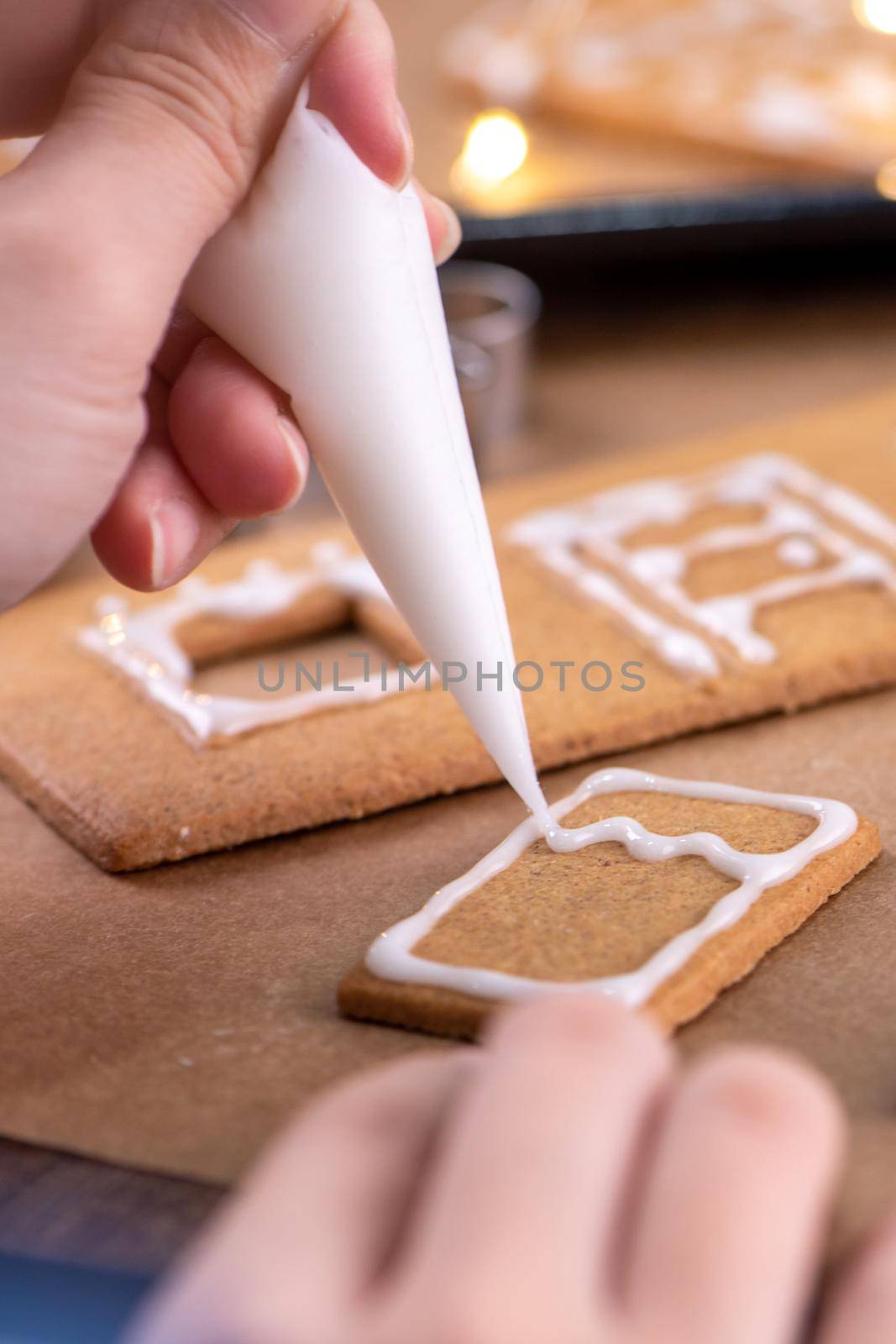 Young woman is decorating Christmas Gingerbread House cookies biscuit at home with frosting topping in icing bag, close up, lifestyle.