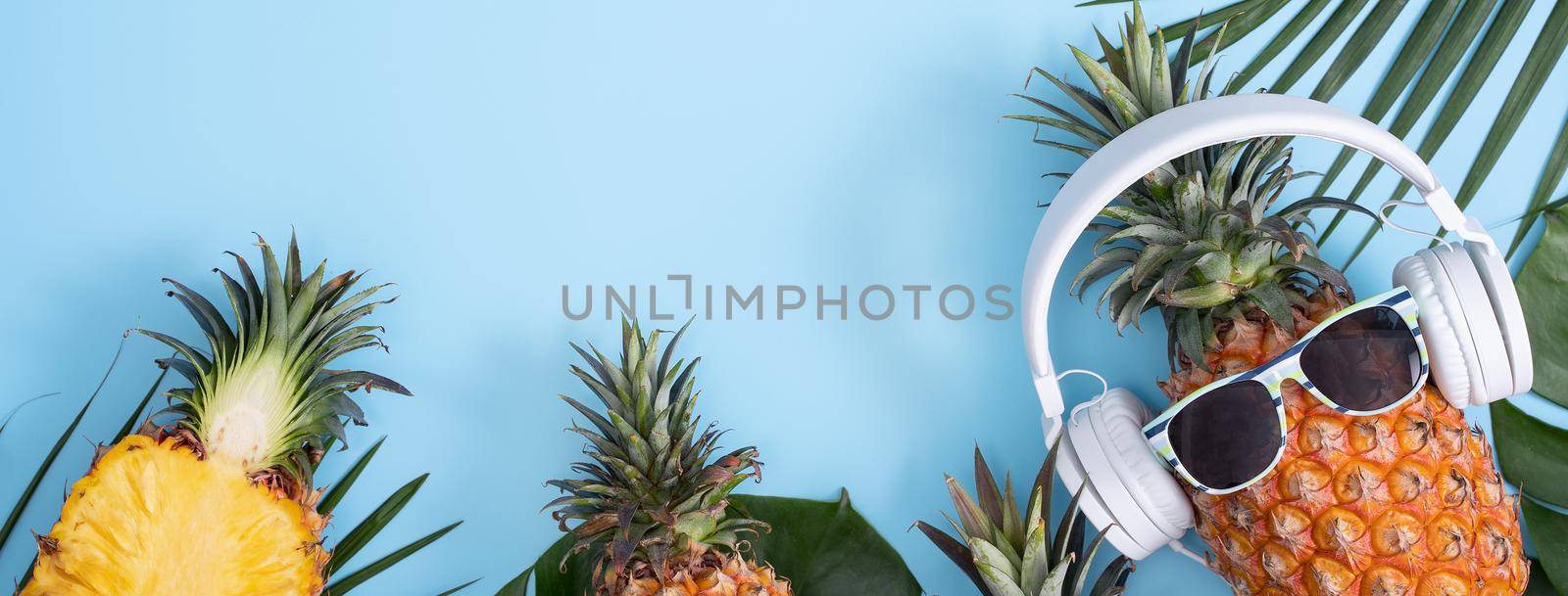 Funny pineapple wearing white headphone, concept of listening music, isolated on colored background with tropical palm leaves, top view, flat lay design.