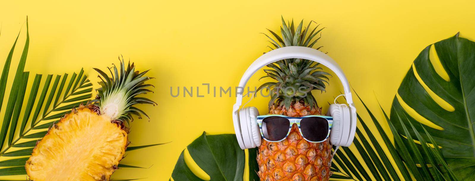 Funny pineapple wearing white headphone, listen music, isolated on yellow background with tropical palm leaves, top view, flat lay design concept.