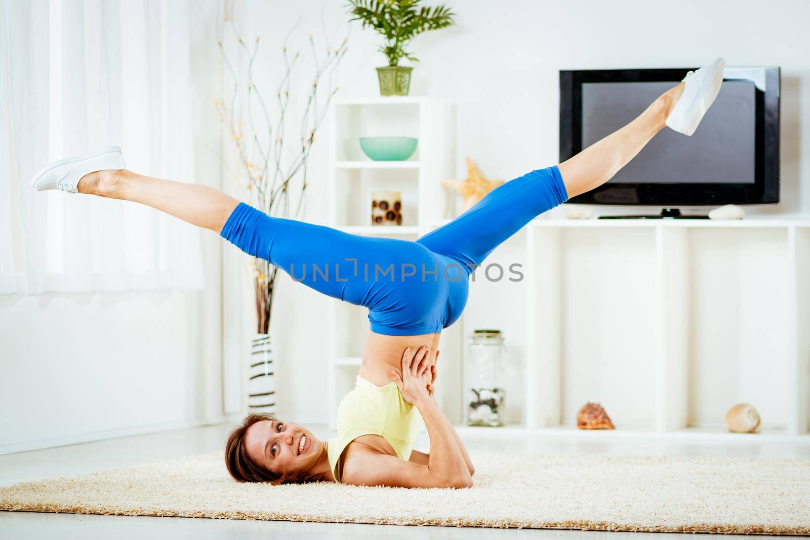 Attractive fitness woman doing exercises and stretching with legs up, in her living room. 