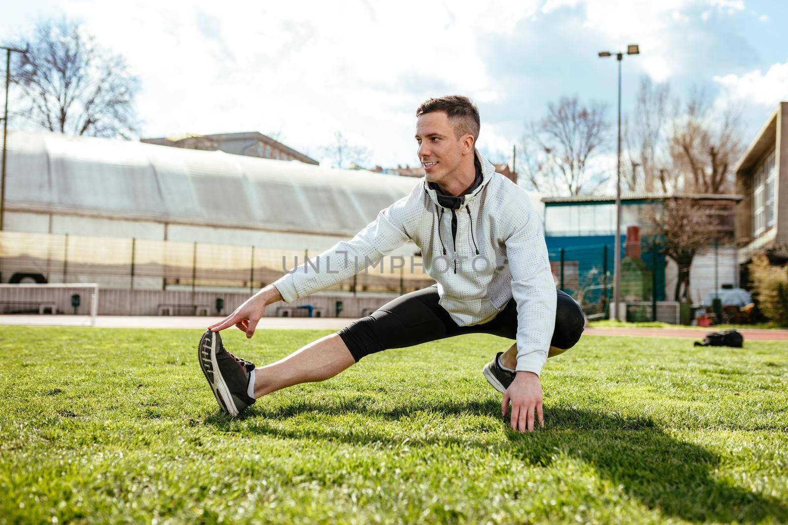 Active young man stretching legs and doing exercises in the public place on empty stadium. 
