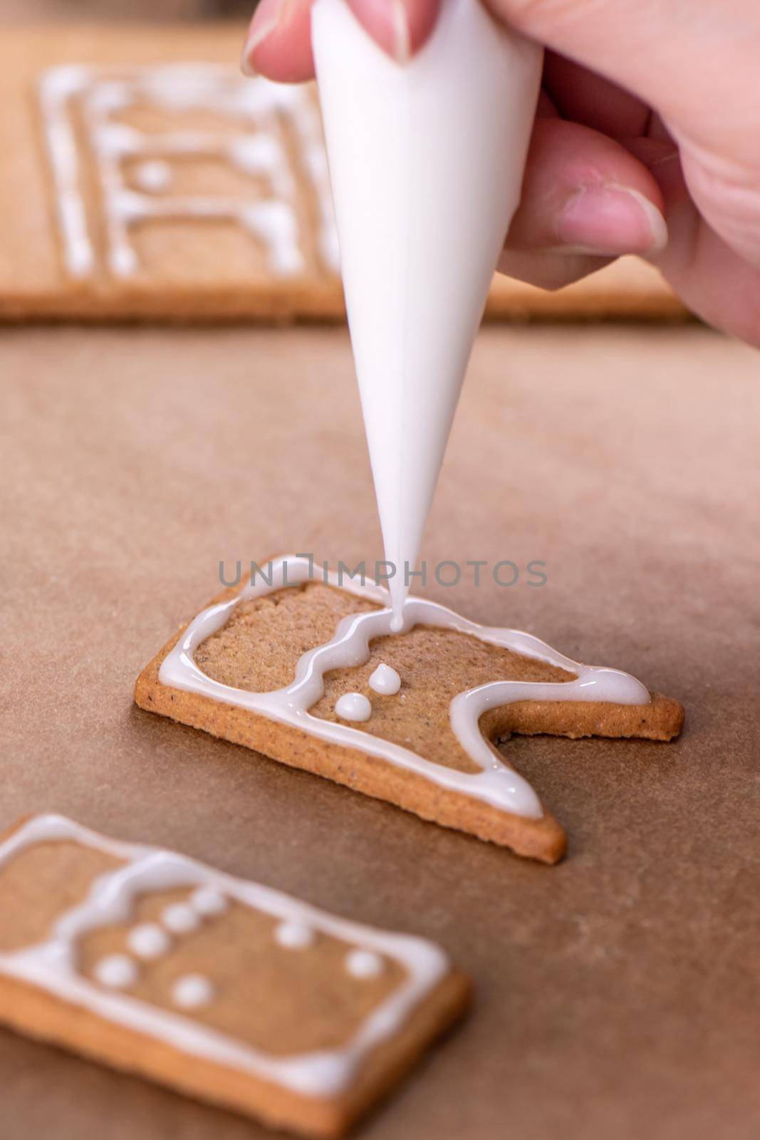 Young woman is decorating Christmas Gingerbread House cookies biscuit at home with frosting topping in icing bag, close up, lifestyle.