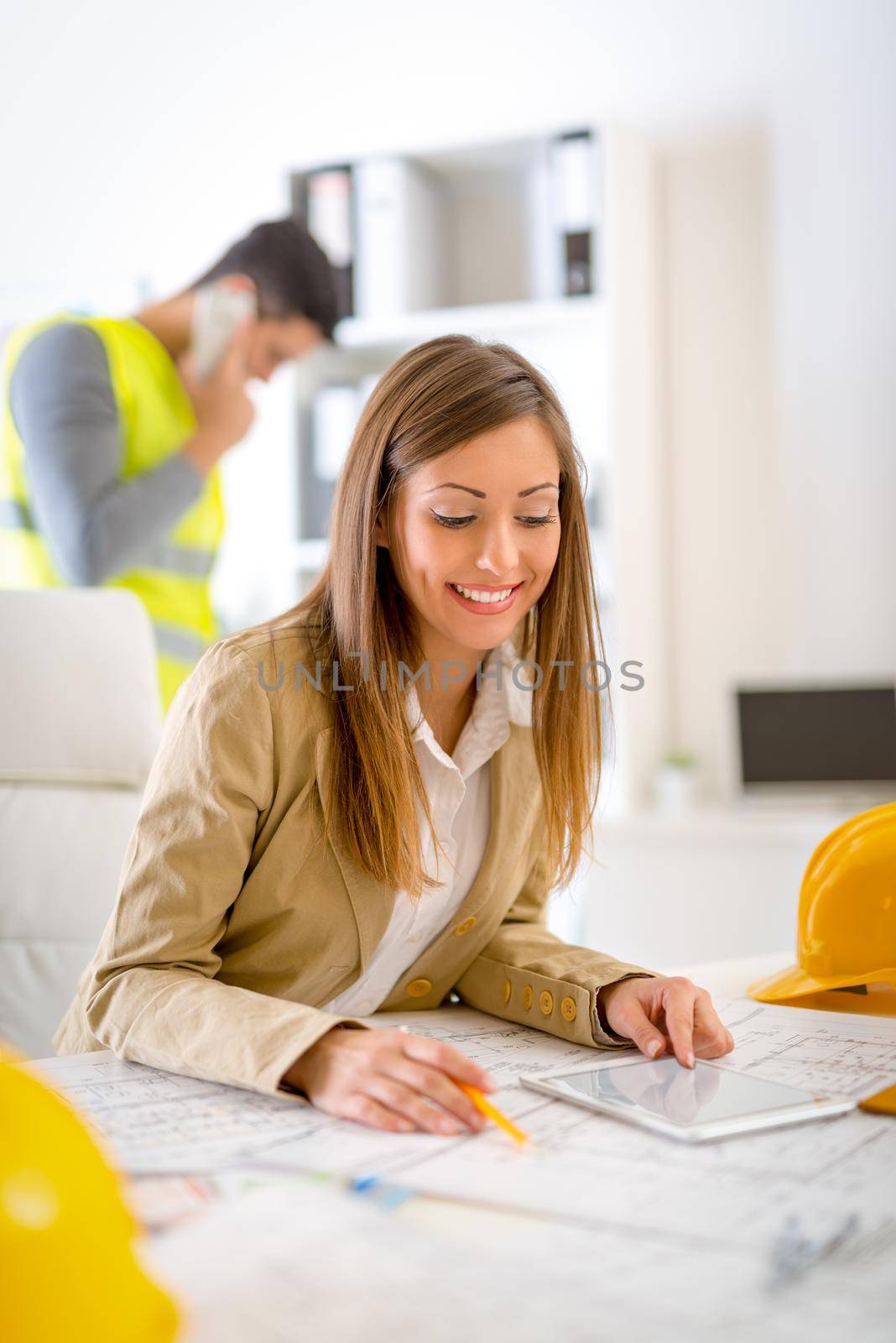 Smiling young woman constructor analyzing blueprint on tablet at desk in office.