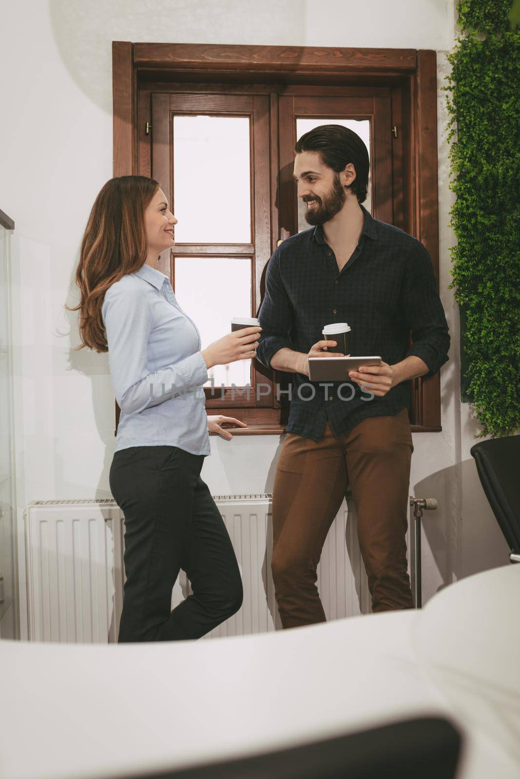 Young smiling entrepreneurs having coffee break and using digital tablet in front of the office window.