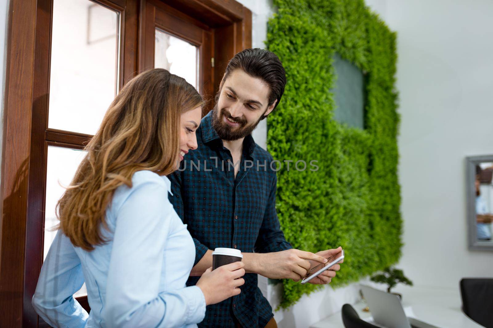 Young smiling entrepreneurs having coffee break and looking something interesting on digital tablet in front of the office window.