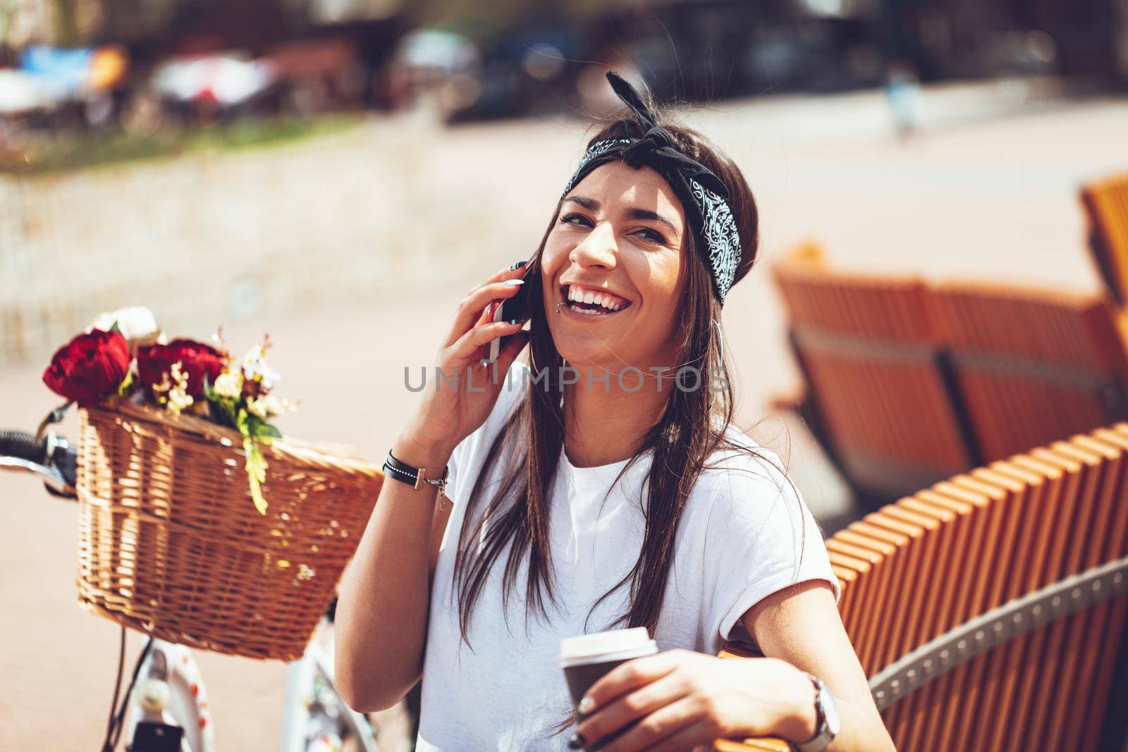 Laughing young woman talking on smartphone, taking coffee break, sitting on a bench on a sunny day, beside the bike with flower basket.