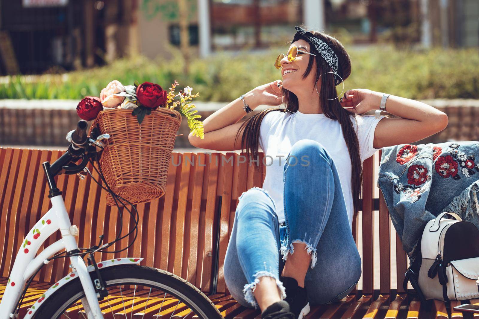 Happy smiling young woman is enjoying in a summer sunny day, sitting  on city bench, beside the bike with flower basket.