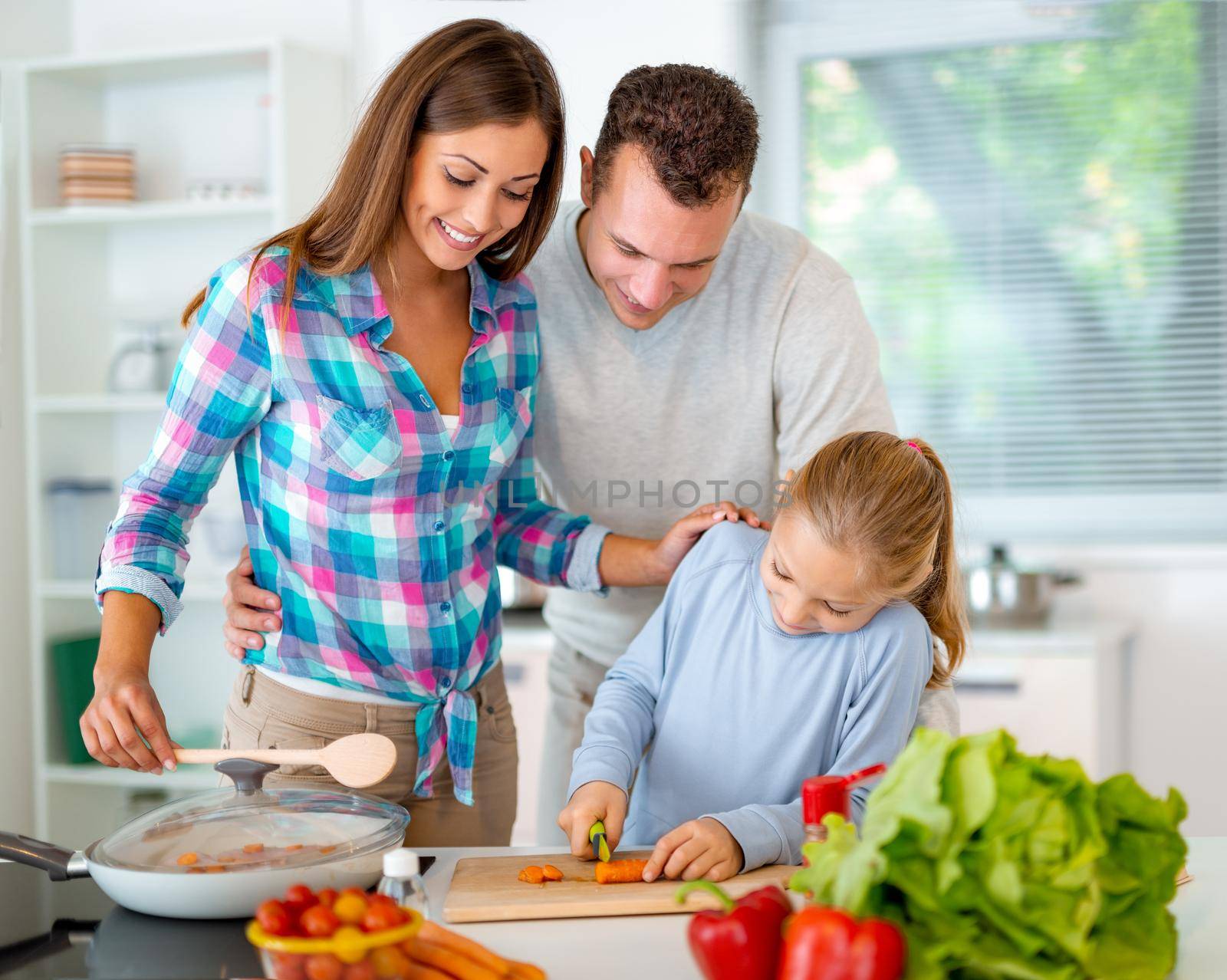 Beautiful young family cooking healthy meal in the domestic kitchen. The little girl learns how to cut the carrot on the kitchen board and her parents supervise her.