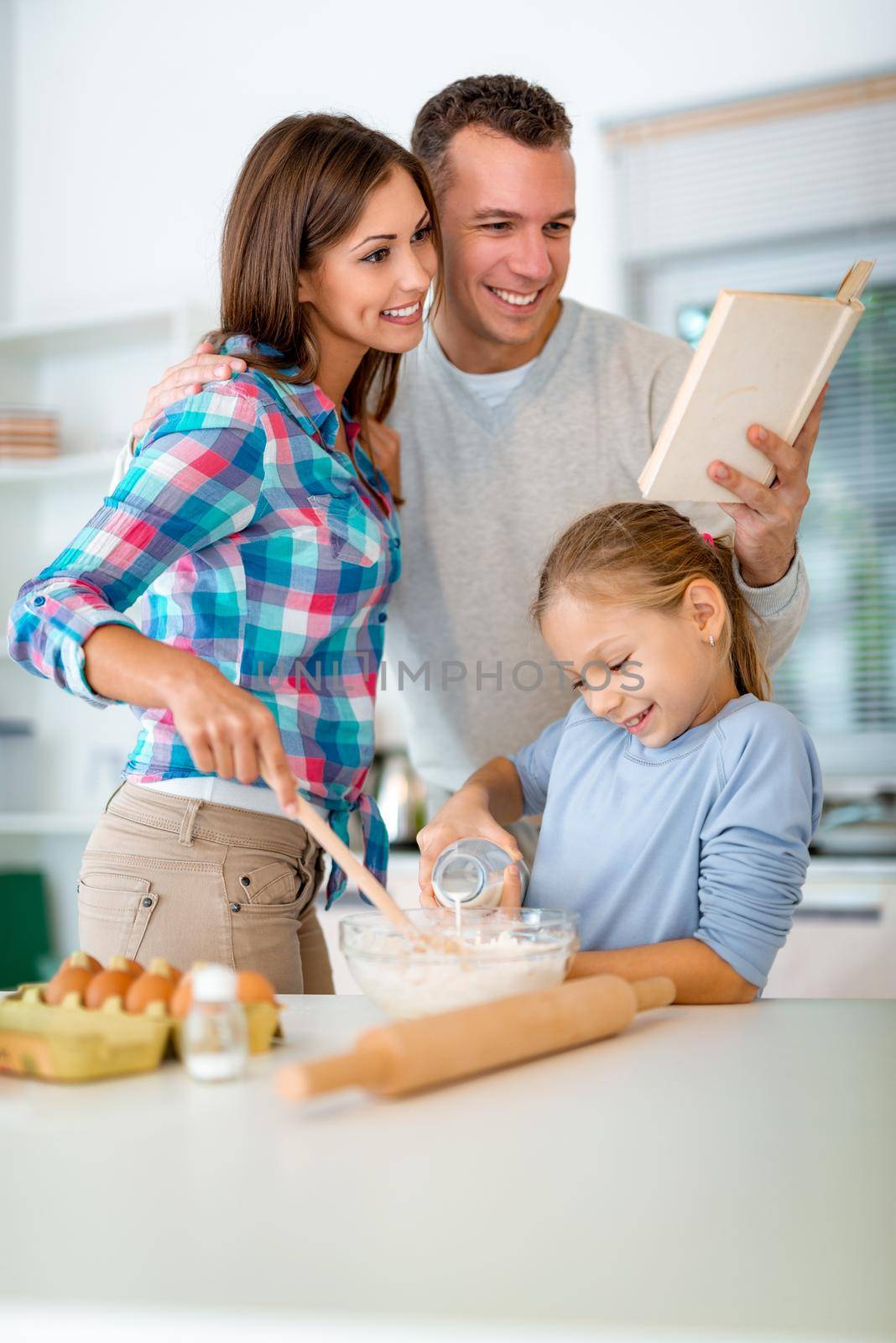 Little girl is pouring the milk in the dish with dough which mother mixing, and father is reading recipe for making cookies in the domestic kitchen.