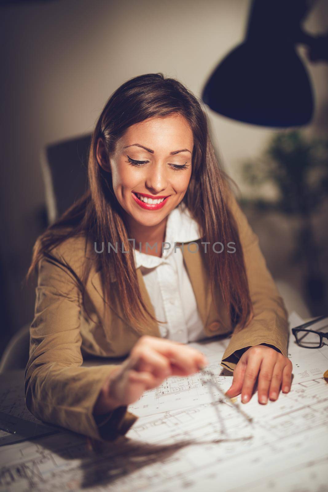 Smiling young woman architect constructor analyzing blueprint at desk in office. She is measuring with drawing divider. Selective focus. 