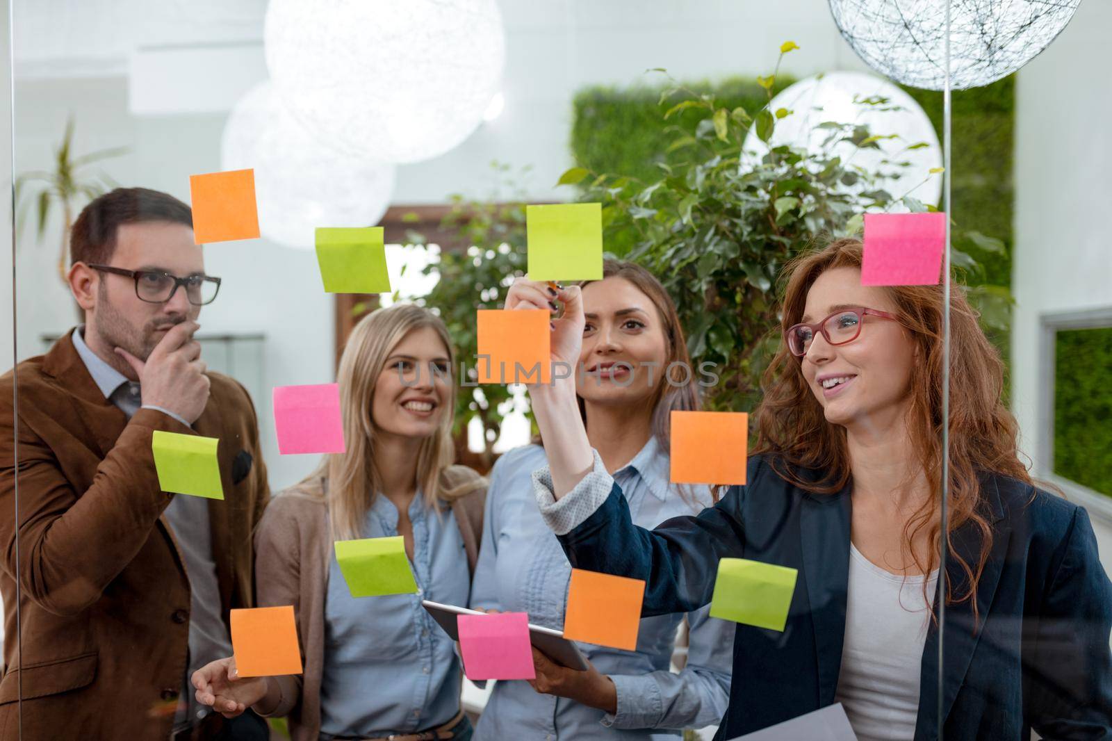 Creative professionals standing and discussing at the office behind glass wall with sticky notes and looking a post it note wall.