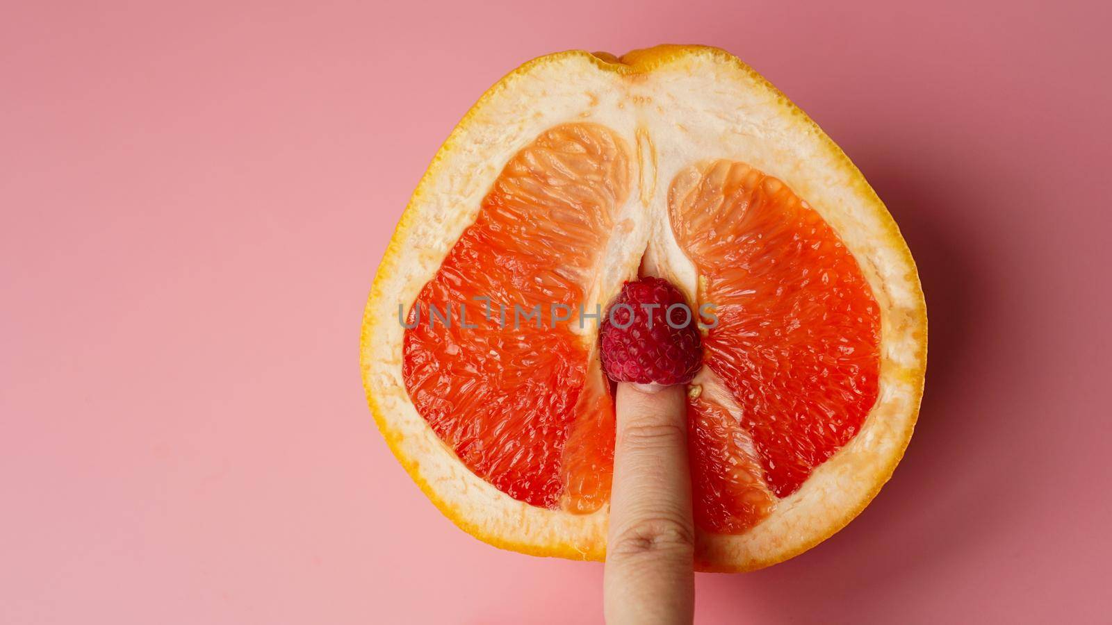 Vagina symbol. Finger with raspberry on grapefruit on pink background. Sex concept.