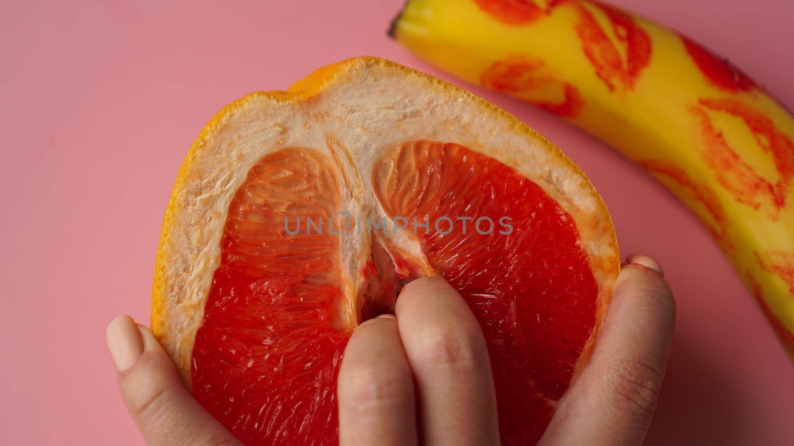 Fingers in grapefruit on pink background. Composition with fresh banana with traces of red lipstick and grapefruit on pink background.