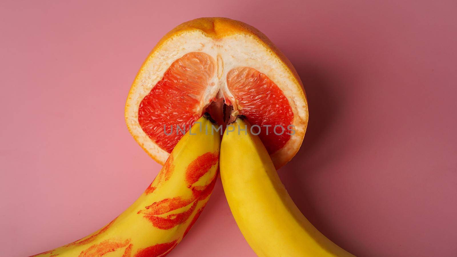 Two bananas and grapefruit on a pink background. There are traces of red lipstick on the banana